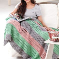 a woman sitting on a couch holding a book and smiling at the camera while wearing a multicolored crocheted blanket