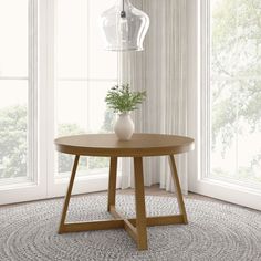 a round wooden table with a plant on it in front of two windows and a rug