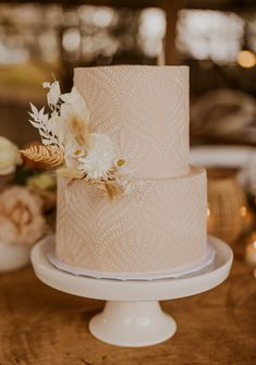 a three tiered wedding cake with white flowers and feathers on the top, sitting on a wooden table