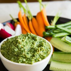 a white bowl filled with green pest next to cucumbers and carrots