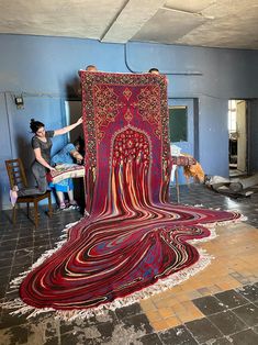 a woman standing next to a large rug on the floor in a room with people sitting around