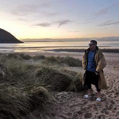 a man standing on top of a sandy beach next to grass and sand dunes at sunset