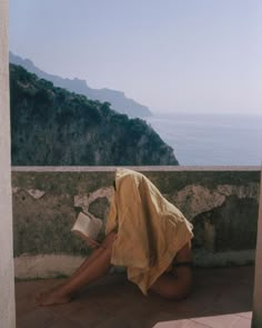 a woman is sitting on the ground reading a book and looking out at the ocean