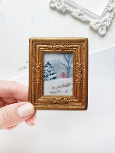 a hand holding up a small gold frame with a snowy scene in the background and a red house