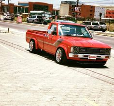 an old red truck is parked on the side of the road with other cars in the background