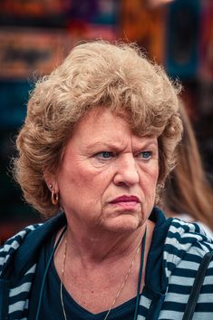 an older woman with blonde hair and blue eyes stares at the camera while standing outside