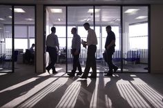 three men are walking in an office lobby with glass doors and sunlight streaming through the windows