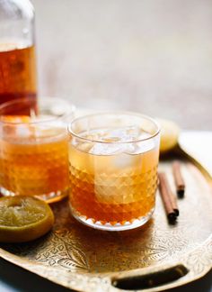 two glasses filled with liquid sitting on top of a tray next to lemons and cinnamon sticks