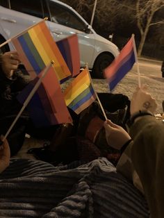 people sitting on the ground holding rainbow flags in front of a white car at night