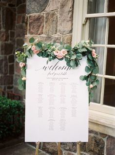 a white sign with greenery and flowers on it sitting in front of a window