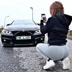 a woman squatting in front of a car taking a photo with her cell phone