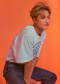 a young man sitting on top of a stool in front of an orange wall wearing a white t - shirt and black pants