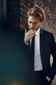 a man in a suit and tie standing next to a brick wall with his hand on his mouth