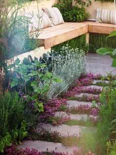 a garden with purple flowers and plants growing on the ground next to a wooden bench