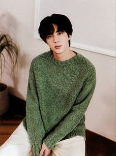 a young man in a green sweater sitting on a wooden table next to a potted plant