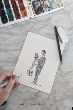 a person holding a paintbrush next to a drawing of a bride and groom on a marble table