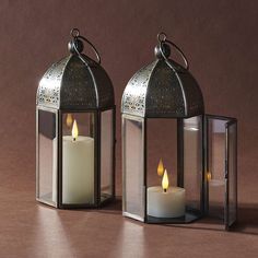 two metal lanterns with lit candles in them on a table next to each other against a brown background