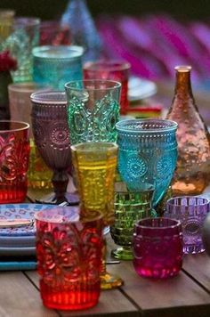 many different colored glasses are sitting on a wooden table with plates and utensils