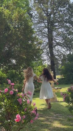 two women in white dresses are walking through the grass with pink flowers and trees behind them