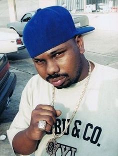 a man wearing a blue hat and holding a chain around his neck while standing in front of parked cars