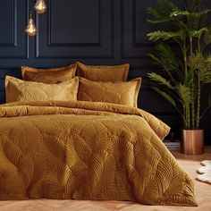 a bed with gold comforter and pillows in a room next to a potted plant