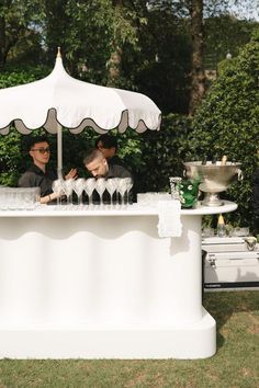 two men sitting under an umbrella at a bar with wine glasses in front of them