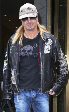 a man with long blonde hair wearing a black shirt and leather jacket standing in front of a building