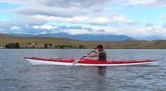 a man in a red and white kayak paddling on the water with mountains in the background