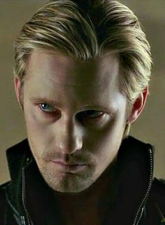 a man with blonde hair and blue eyes looks at the camera while wearing a black jacket