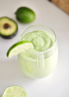 an avocado smoothie in a glass next to sliced limes on a white surface