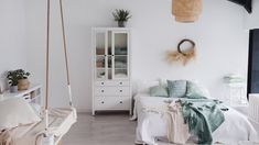 a bedroom with white walls and wood flooring has a bed, hanging planter, and chair