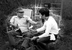 two men sitting on lawn chairs in the grass talking to each other with one holding a pen