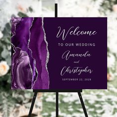 a purple and white wedding welcome card on a easel with flowers in the background