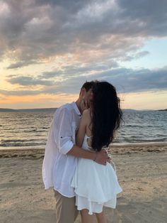 a man and woman kissing on the beach at sunset with clouds in the sky behind them