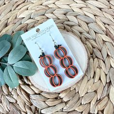 an orange and black pair of earrings sitting on top of a woven basket next to a plant