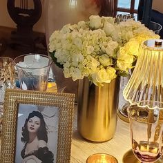 a table topped with gold vases filled with white flowers and pictures next to candles