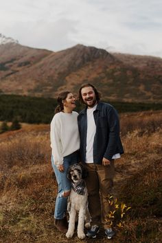 a man and woman standing next to a dog on top of a grass covered field