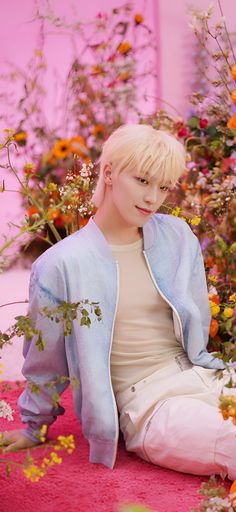 a young man sitting on the ground in front of flowers and plants, wearing a blue jacket