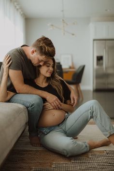 a pregnant couple cuddle on the floor in their living room while looking at each other