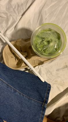 a bowl of green liquid sitting on top of a bed next to a pair of jeans