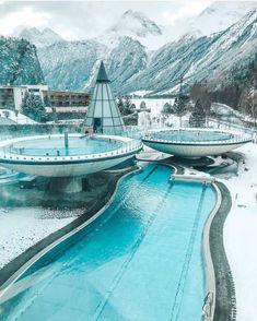 an outdoor swimming pool surrounded by snow covered mountains and trees with buildings in the background