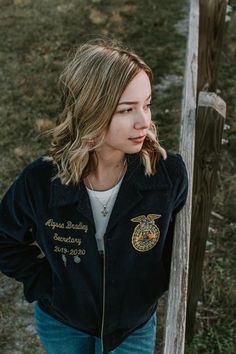 a woman standing next to a wooden fence wearing a black jacket with embroidered words on it