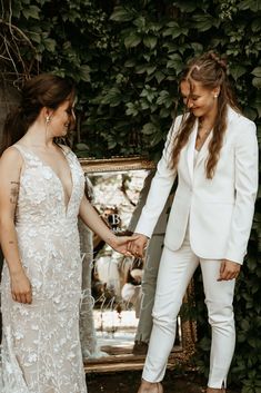 two women standing next to each other in front of a mirror and ivy covered wall