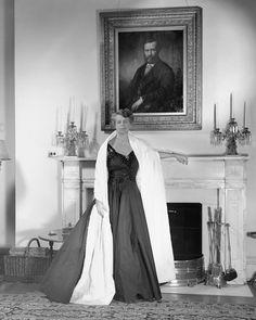 an old black and white photo of a woman in a formal dress standing in front of a fireplace