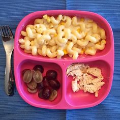 a pink plate with pasta, grapes and meat on it next to a silver fork