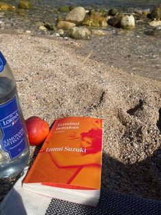 a bottle of water sitting next to an orange book on the beach with rocks in the background