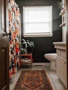 a white toilet sitting next to a bath tub in a bathroom under a window with a rug on the floor