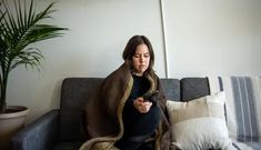 a woman sitting on a couch looking at her cell phone while wrapped in a blanket