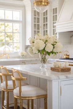 a white kitchen with gold accents and flowers in a vase on the island countertop