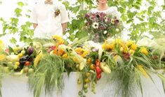 two people standing in front of a table filled with flowers and vegetables, one holding a bouquet